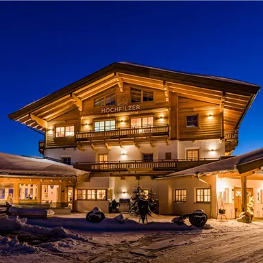 Ein charmantes Holzhaus in den Bergen, beleuchtet von warmem Licht in der Dämmerung. Umgeben von Schnee und einer ruhigen Winterlandschaft.
