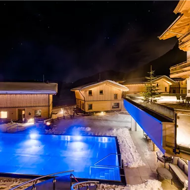 Eine beleuchtete Terrasse mit einem Pool in der Nacht. Umgeben von Chalets und schneebedeckter Landschaft.