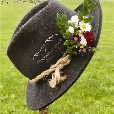 Ein grauer Hut mit einer Blumenverzierung steht auf einem Stock in einer Wiese. Im Hintergrund sind grüne Felder und Berge zu sehen.