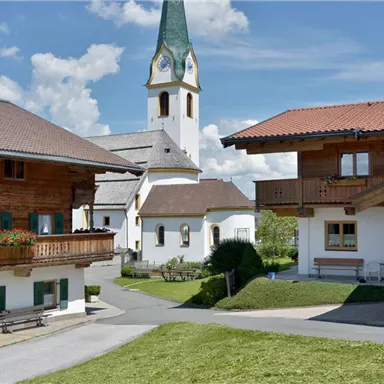 Ein malerisches Dorf mit traditionellen Holzhäusern und einer Kirche im Hintergrund. Der Himmel ist klar und blau, was eine ruhige Atmosphäre schafft.