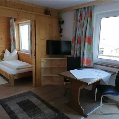 A cozy room with wooden walls and a window that lets in a lot of light. The room contains a bed, a television, a table, and a chair.