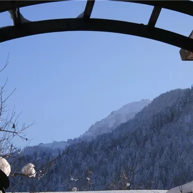Eine verschneite Berglandschaft unter einem klaren blauen Himmel. Im Vordergrund ist ein architektonischer Bogen zu sehen.
