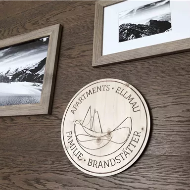 Ein Holzschild mit der Aufschrift "Apartments Ellmau Familie Brandstätter" hängt an einer Holzwand. Daneben befinden sich zwei eingerahmte Schwarzweißbilder von Landschaften.