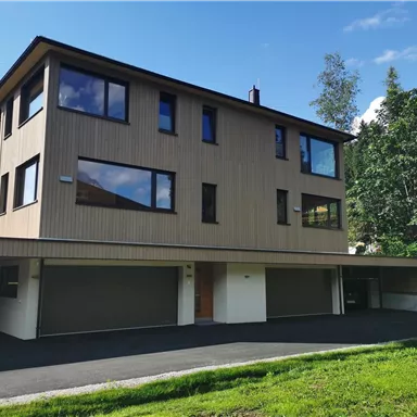 A modern house with a wooden facade and large windows. It is situated in a green environment with a well-maintained garden.
