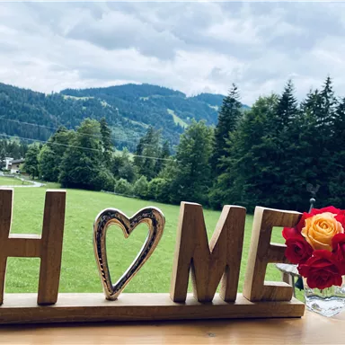 A wooden decoration with the word "HOME" stands out in the foreground. In the background, a green landscape with mountains and trees can be seen.