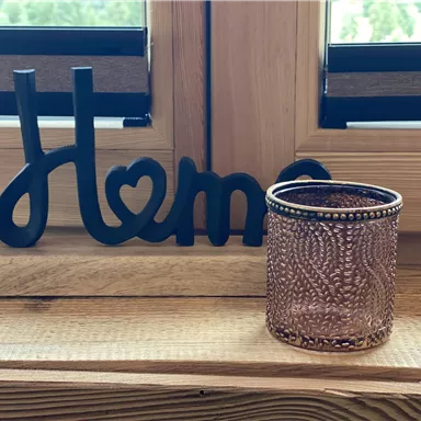A decorative "Home" sign made of wood stands next to an elegant glass container on a wooden windowsill. The scene radiates coziness and a sense of home.