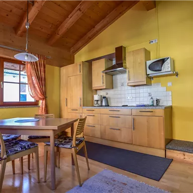 Eine gemütliche Küche mit Holzmöbeln und einer Essecke. Die Wände sind gelb gestrichen und große Fenster sorgen für viel Tageslicht.
