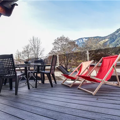 Eine Terrasse mit einem Tisch und vier Stühlen, darunter zwei Liegestühle in Rot. Im Hintergrund sind Bäume und Berge zu sehen, unter einem klaren Himmel.