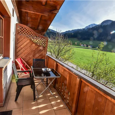 Ein gemütlicher Balkon mit einem Tisch und Stühlen bietet einen Blick auf grüne Wiesen und Berge im Hintergrund. Die Sonne scheint auf die entspannende Umgebung.