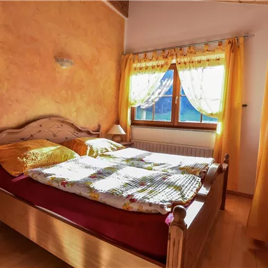 Ein gemütliches Schlafzimmer mit einem großen Bett und bunten Bettwäsche. Helle Vorhänge lassen viel Tageslicht herein.