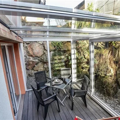 Ein moderner Wintergarten mit Glaswänden und einem Holzboden. In der Mitte stehen ein Tisch und Stühle, umgeben von einer natürlichen Steinwand.