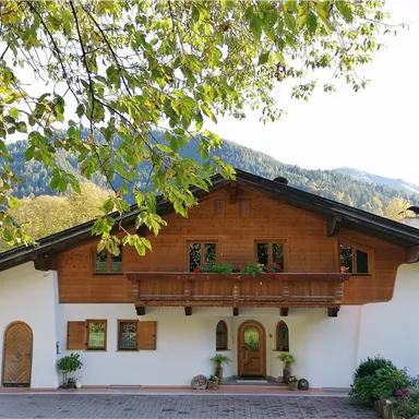 A traditional Alpine house with wooden siding and a blooming balcony. Surrounded by green trees and gentle hills.