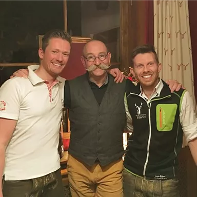 Drei Männer posieren in traditioneller Kleidung vor einem Fenster. Sie lächeln und scheinen eine gesellige Atmosphäre zu genießen.