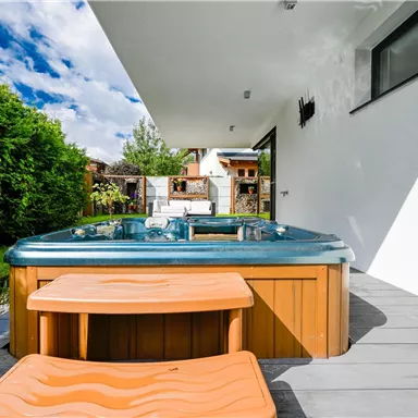 Ein gemütlicher Außenbereich mit einem Whirlpool und einer Holzbank. Die Umgebung ist grün und hell beleuchtet von der Sonne.