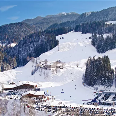 Eine malerische Winterlandschaft mit schneebedeckten Bergen und einem Skigebiet. Im Vordergrund sind ein Parkbereich und einige Gebäude zu sehen.