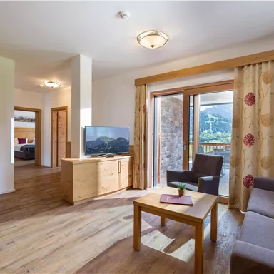 A bright and modern apartment with wooden flooring and large windows. The room features a cozy sofa, a table, and a television.