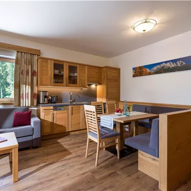 A cozy holiday apartment with modern wooden interior. The living area includes a kitchenette, a dining area, and plenty of daylight through the large windows.