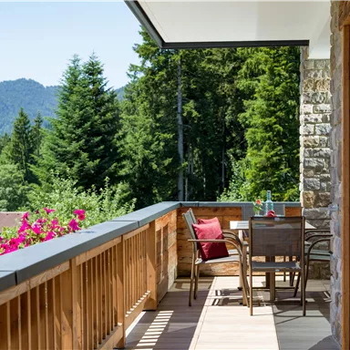 A cozy balcony with wooden furniture and beautiful plants. In the background, green trees and mountains rise.