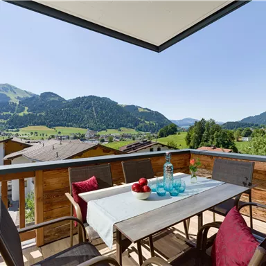 A beautiful balcony with a view of the mountains and green forests. The table is set with glasses and a decorative bowl.