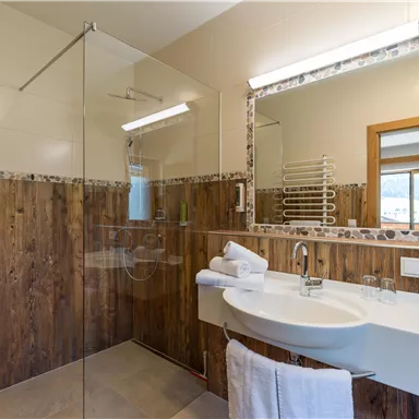 A modern bathroom with wood paneling and a large mirror. The shower has a glass partition and fresh towels are ready.