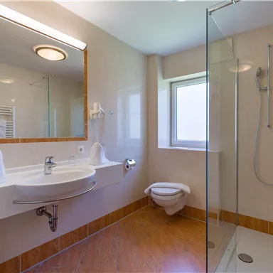 A modern bathroom with a glass shower and a sink. Many bright surfaces and a large window create an open ambiance.