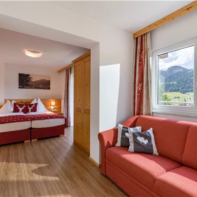 A bright room with a red sofa and a double bed. In the background, mountains and a clear sky are visible.