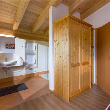 A bright room with wooden ceilings and a bathroom area. There is a wooden wardrobe and a clear access to another room.