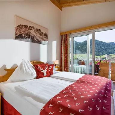 A cozy hotel room with a large double bed and a beautiful view of the mountains. The balcony is decorated with flowers and offers an inviting view outside.