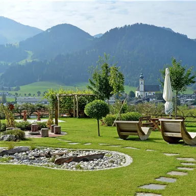 Garden area of Hotel Alpenpanorama in the Wilder Kaiser holiday region with sun loungers, circular bench, and panoramic view of the Tyrolean Alps.