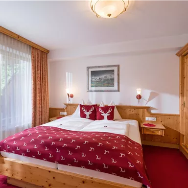 A cozy hotel room with a large double bed and rustic wooden furniture. The windows are adorned with light curtains, and the carpet is red.