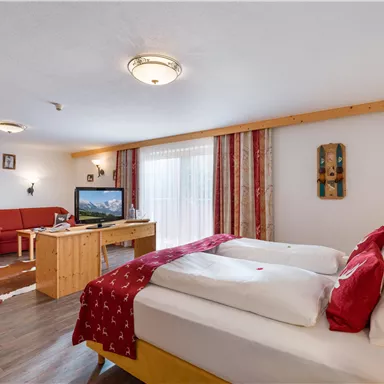 A cozy room with two beds, a table, and a red sofa. Large windows provide plenty of daylight.