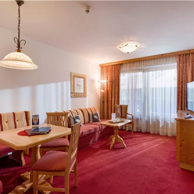 A cozy living room scene with a red carpet, a dining table, and a seating area. Large windows let in plenty of light.