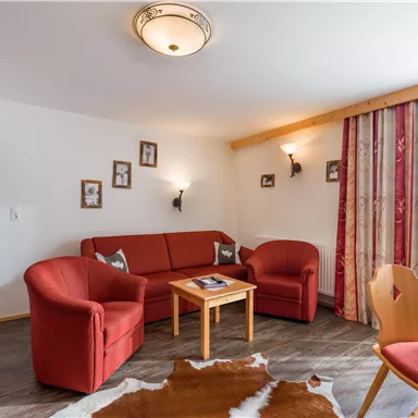 A cozy living room with a red sofa set and a wooden table. Pictures hang on the wall and there are large windows with curtains.