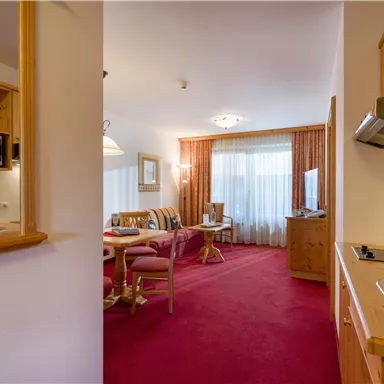 A cozy living room with a red carpet and large windows. The kitchen is open and features wooden cabinets and modern appliances.