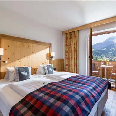 A cozy bedroom with a large bed and wooden decor. In the background, a balcony with a view of the mountains can be seen.