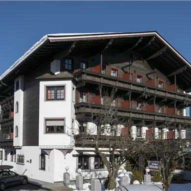 Ein traditionelles Alpenhotel mit mehreren Balkonen und einem großen Dach. Schnee liegt rund um das Gebäude, und die Berge sind im Hintergrund sichtbar.