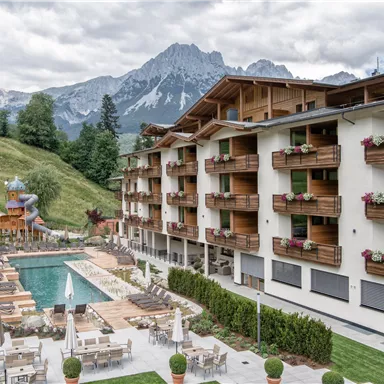A modern hotel with balconies and a swimming pool. In the background, majestic mountains and lush landscapes can be seen.