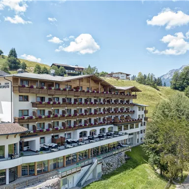 A modern hotel with several balconies, surrounded by green hills and mountains. The sky is clear and sunny.