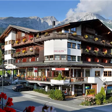 A charming hotel in the Alps with wooden decorations and flower-filled balconies. In the background, mountains and a green landscape are visible.