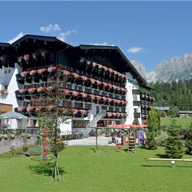 Ein malerisches Hotelgebäude mit Balkonen, die mit Blumen geschmückt sind. Im Hintergrund sind grüne Wiesen und beeindruckende Berge zu sehen.