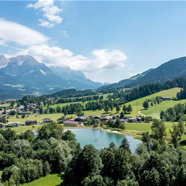 Eine idyllische Landschaft mit einem klaren See und üppigen grünen Wiesen. Im Hintergrund erheben sich majestätische Berge unter einem blauen Himmel.