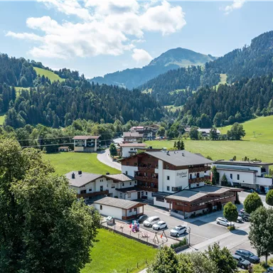 Eine malerische Landschaft mit grünen Wiesen und Bergen im Hintergrund. Im Vordergrund sind mehrere Gebäude und parkende Autos zu sehen.