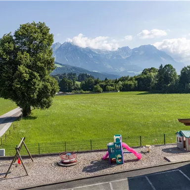 Eine grüne Wiese mit Bergen im Hintergrund. Im Vordergrund gibt es einen Spielplatz mit verschiedenen Spielgeräten.