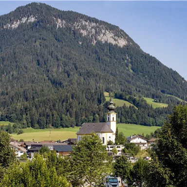 Ein malerisches Dorf mit einer Kirche im Vordergrund und majestätischen Bergen im Hintergrund. Die grüne Landschaft ist von Bäumen umgeben.