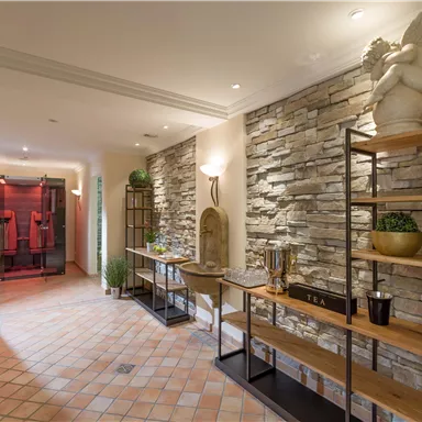 Ein eleganter Flur mit Steinmauern und warmem Licht. Links steht ein Regal mit Pflanzen und dekorativen Objekten.