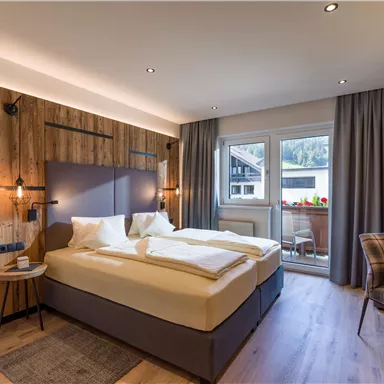 Ein modernes Hotelzimmer mit einem Doppelbett und stilvollem Holzdekor. Große Fenster bieten Blick auf einen Balkon mit Pflanzen.