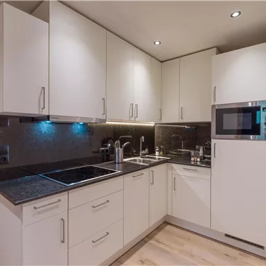 Eine moderne Küche mit weißen Schränken und schwarzer Arbeitsplatte. Sie ist gut ausgestattet mit einem Waschbecken, Kochfeld und Mikrowelle.