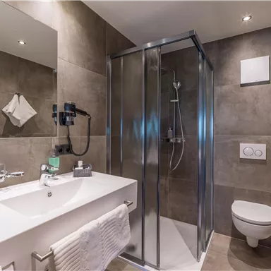 Ein modernes Badezimmer mit einer Dusche, einem Waschbecken und einer Toilette. Die Wände sind in neutralen Tönen gehalten und die Ausstattung ist minimalistisch.