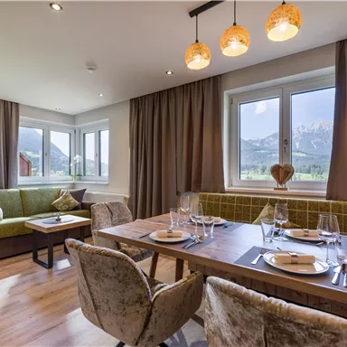 Eine stilvolle Wohnküche mit einem Esstisch und bequemen Sesseln. Große Fenster bieten einen Blick auf die Berge und viel Tageslicht.