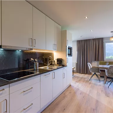 Eine moderne Küche mit weißen Schränken und einer dunklen Arbeitsplatte. Im Hintergrund befindet sich ein gemütlicher Essbereich mit einem Tisch und Stühlen.